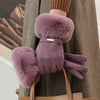 A close-up of a person wearing plush, mauve wrist gloves over a brown coat and holding a tan leather handbag.