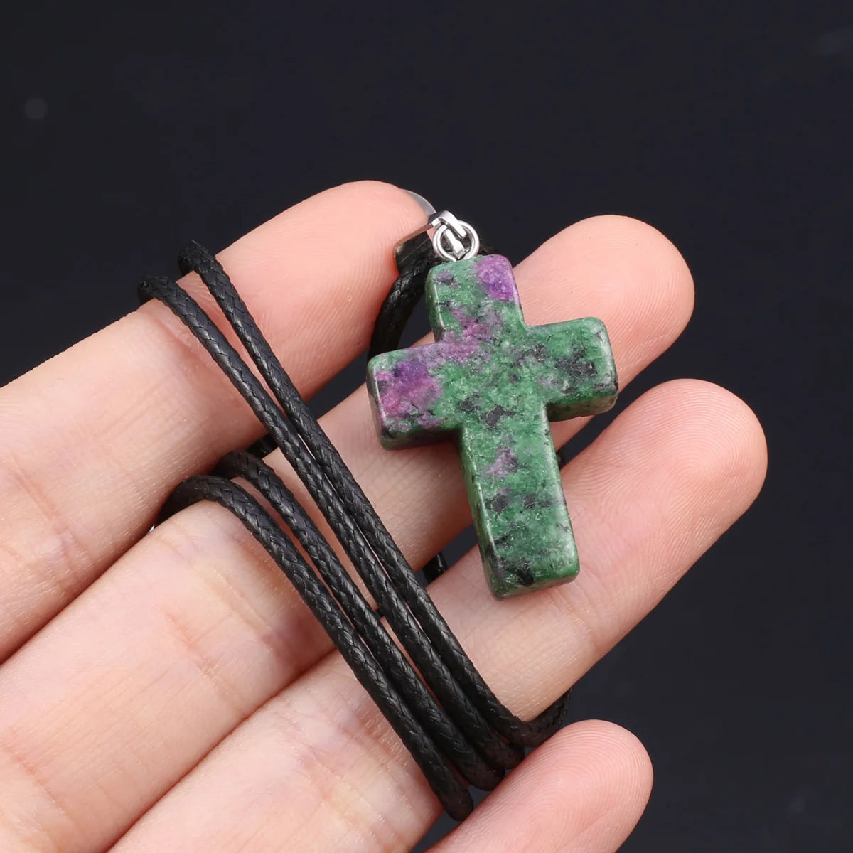 Eine Hand hält eine schwarze Schnurkette mit einem grüne und violette Tigerauge-Agat-Kreuzanhänger.