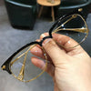 A hand holds a pair of black and gold metal half-frame reading glasses against a blurred background.