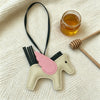 A beige horse charm with pink accents hangs from a black cord next to a small glass jar of honey.