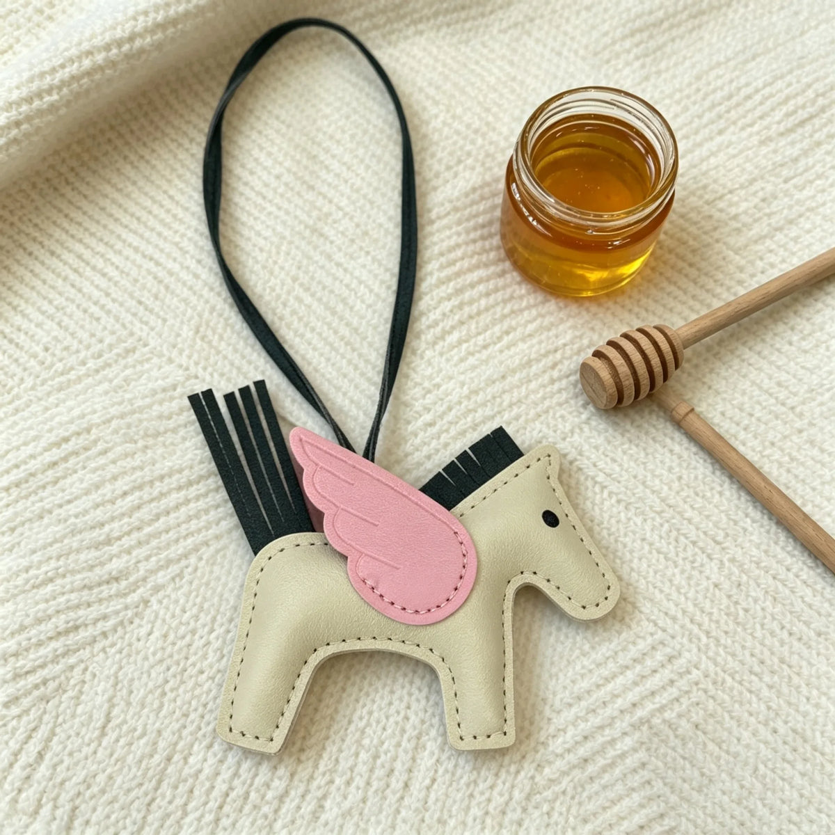 A beige horse charm with pink accents hangs from a black cord next to a small glass jar of honey.