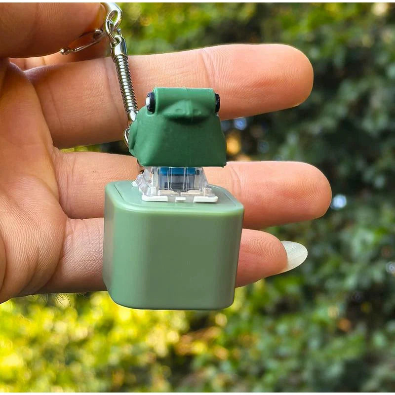 A hand holds a green fidget frog keychain with a clear top piece against a blurred green background.