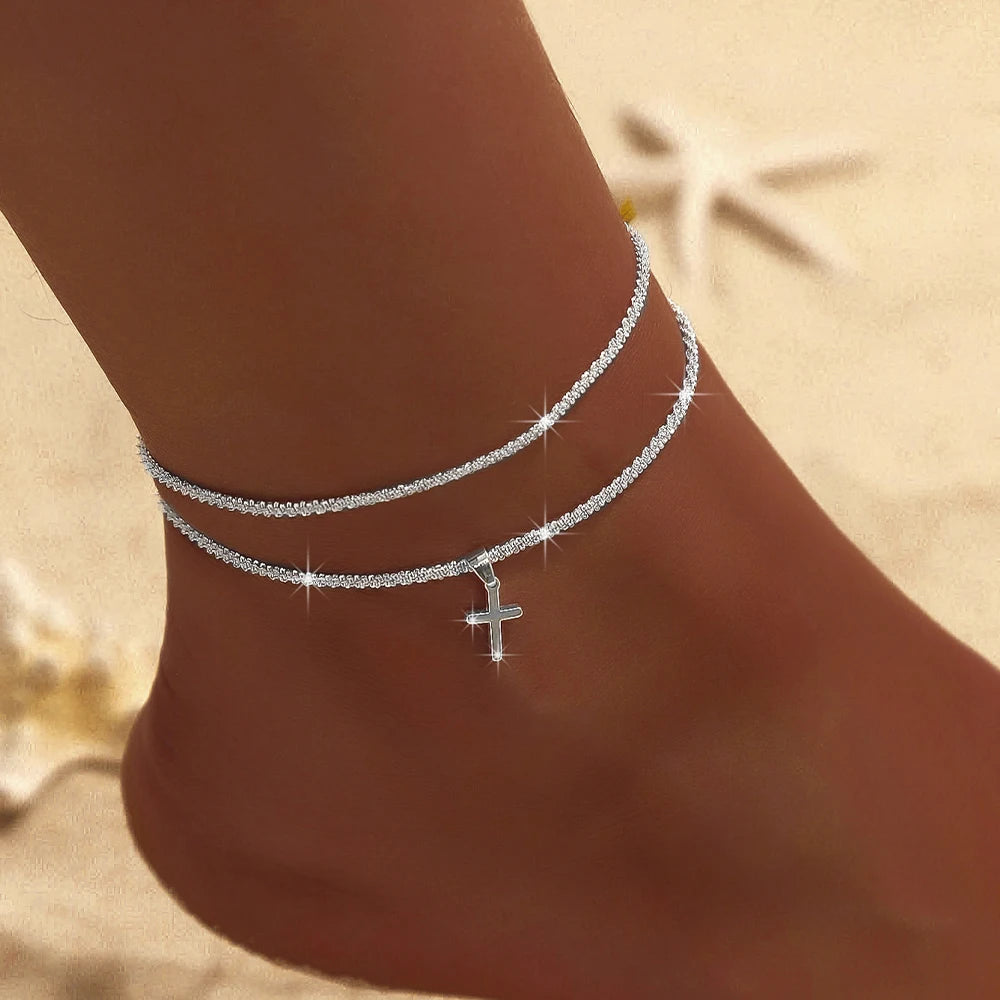 A close-up of a delicate silver-plated CZ cross anklet worn on a person's ankle against a sandy background.