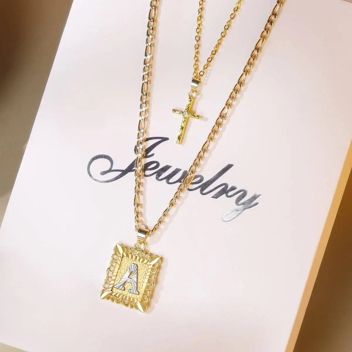 Collier doré avec pendentif lettre A et charm croix, présenté sur une carte rose pâle.