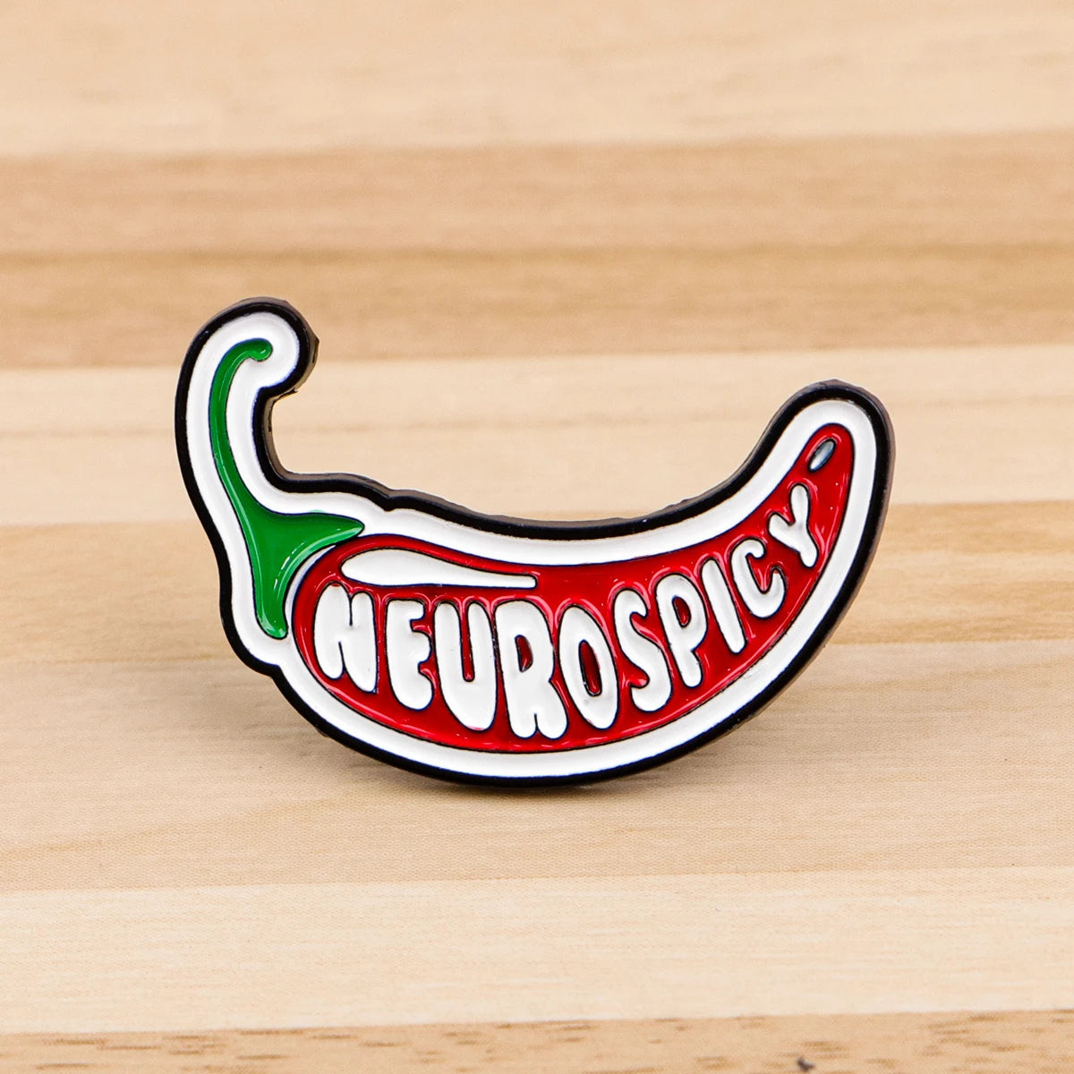 A red chili pepper shaped enamel pin with the word NEUROSPICY on it, resting on a wooden surface.