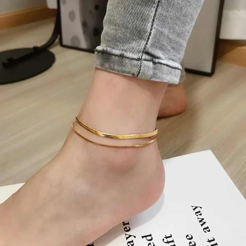 A close-up of a gold double snake anklet worn on a person's ankle against a light background.