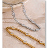 Two necklaces are displayed on a light surface: one silver stainless steel with clear CZ stones and one gold-toned chain with