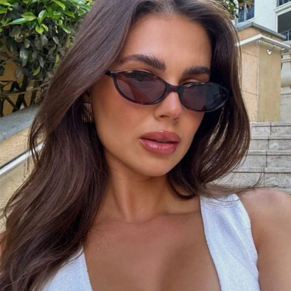 امرأة بشعر داكن طويل ترتدي نظارات شمسية بإطار بيضاوي قديم وبلوزة بيضاء.