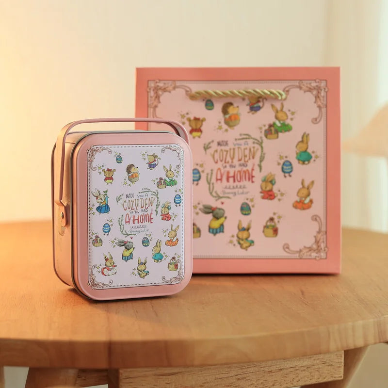 A pink tinplate candy box with a carrying loop sits in front of a framed illustration featuring cute animal characters.