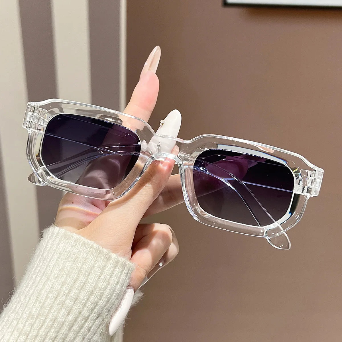 A hand holds a pair of clear acetate retro square sunglasses with dark lenses against a neutral background.