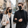 A young man and woman standing outdoors wearing black face masks and coats.