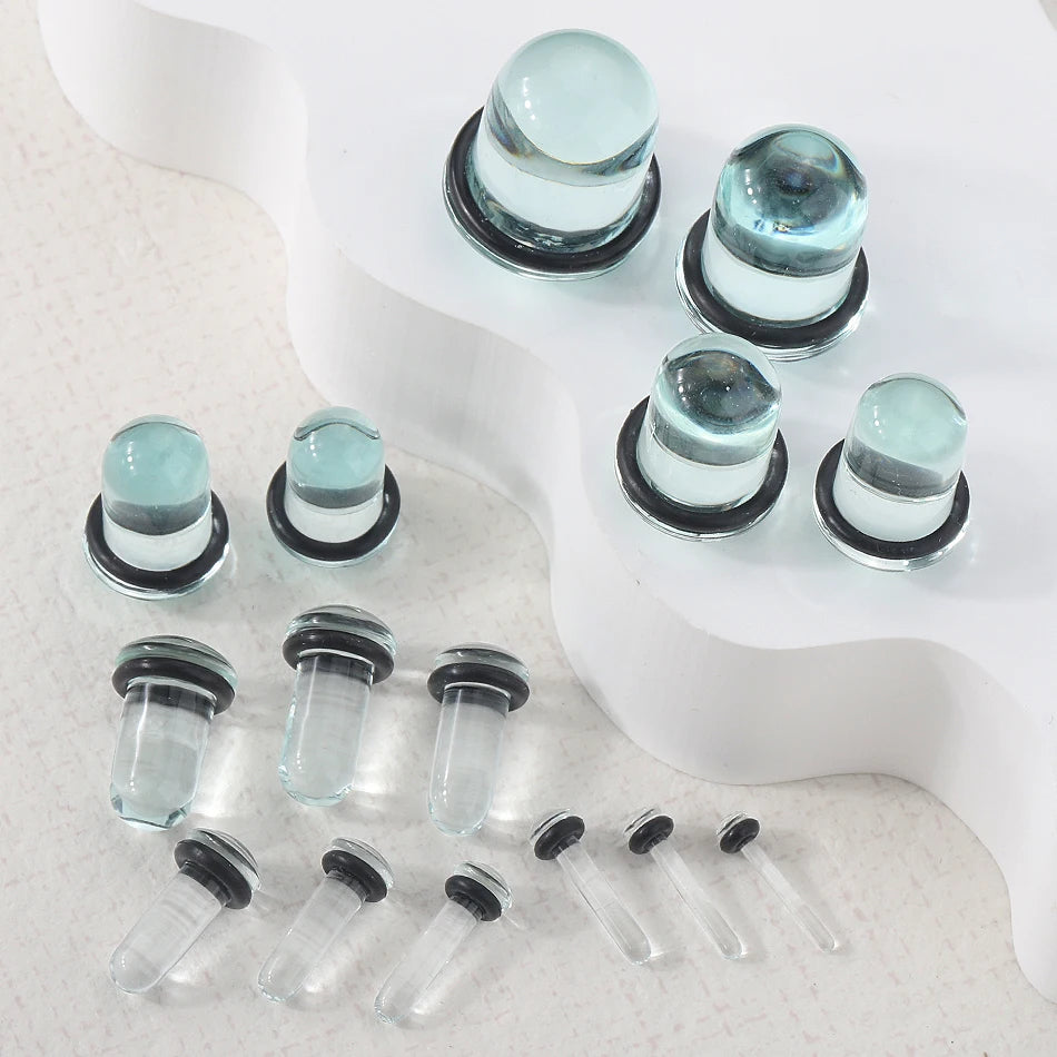 A collection of clear glass ear stretching plugs and tunnels with silver bases displayed on a white surface.