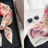 A floral print silk scarf in peach and pink tones is displayed on a dark tray.