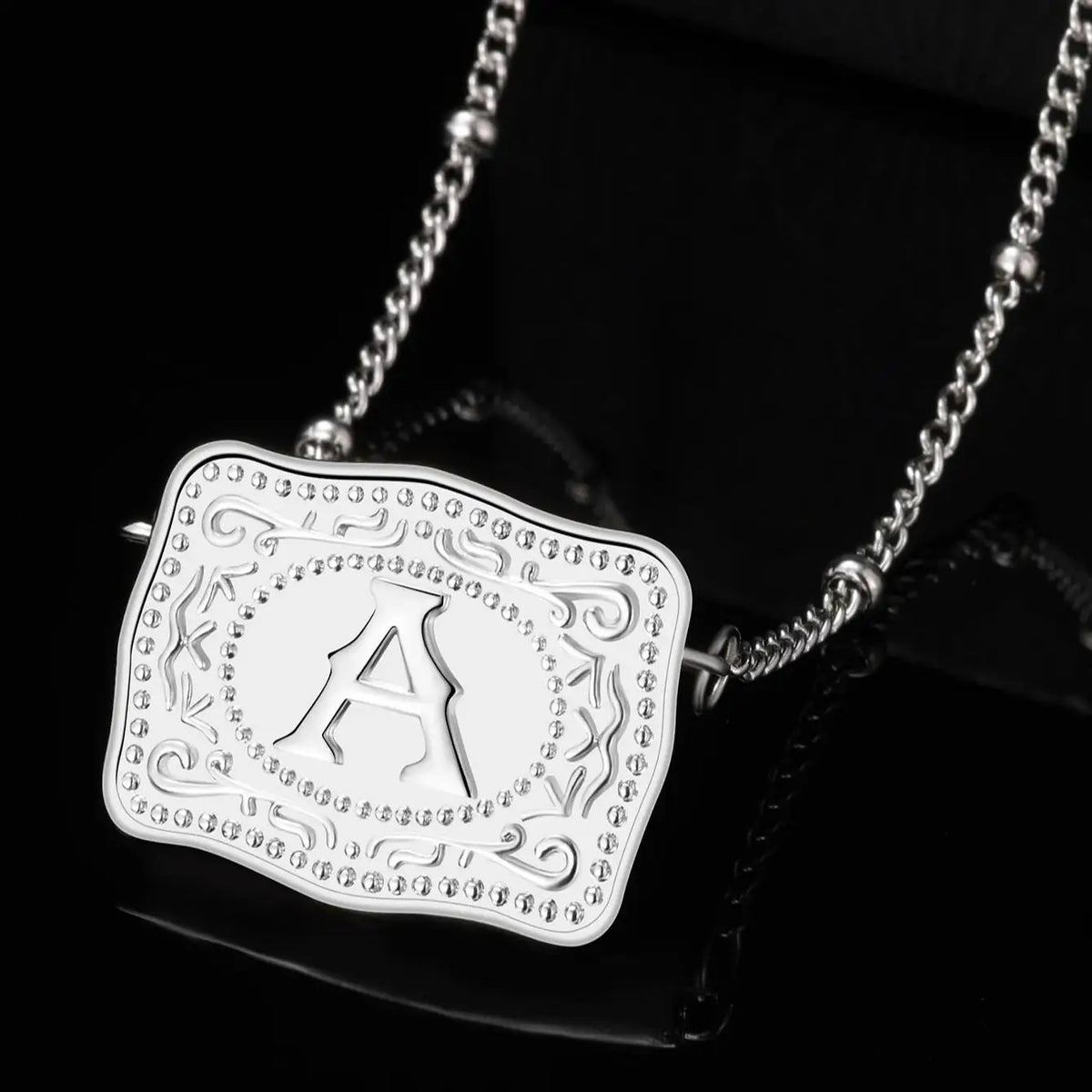 Silver chain necklace with a white initial letter pendant on black fabric.