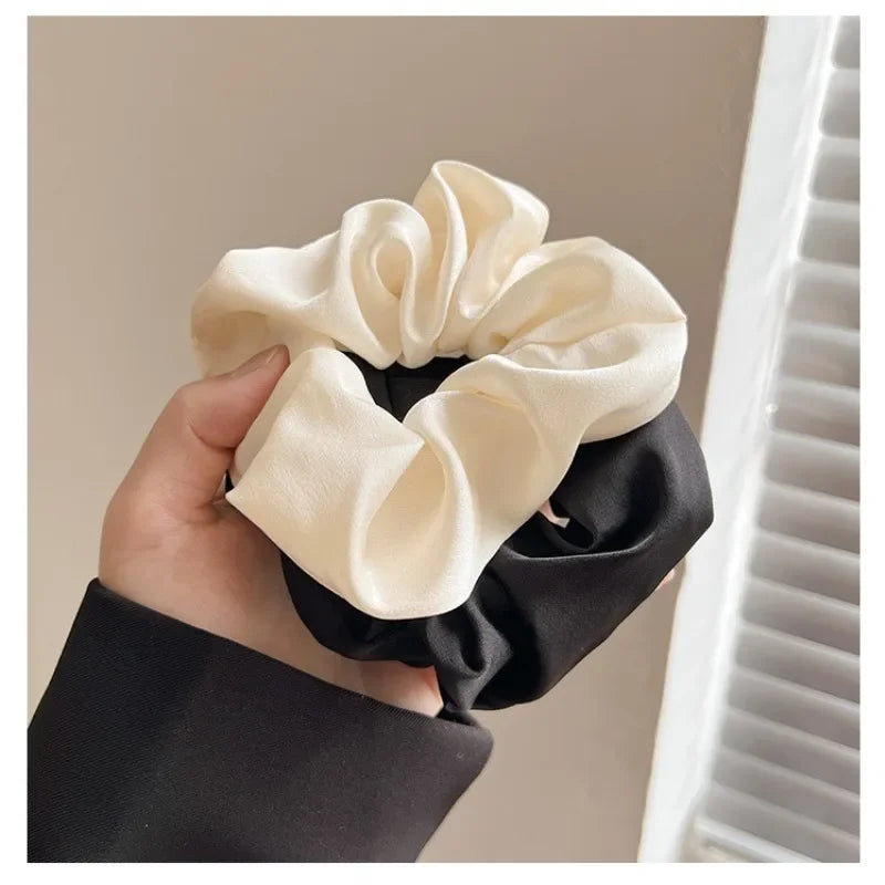 A hand holds a black silk scrunchie hair tie with a cream-colored fabric bow against a white background.