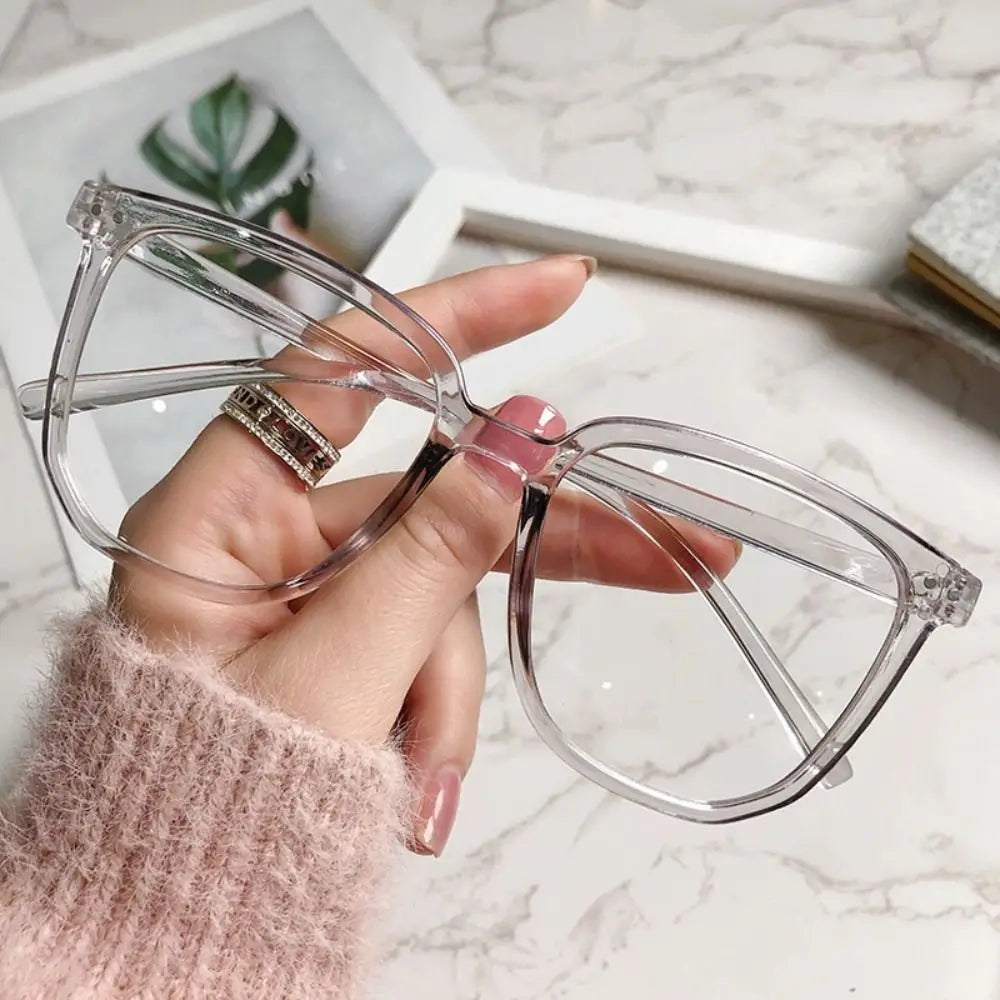 A hand holds a pair of clear, large square anti-blue light spectacles against a marble background.