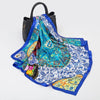A luxury silk square scarf featuring intricate blue, white, and yellow floral patterns tied with a black strap.