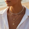 A close-up of a woman wearing a layered gold necklace featuring white pearls and a starfish pendant.