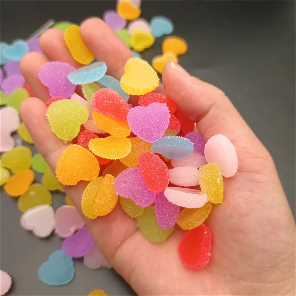 A hand holds a pile of colorful heart-shaped resin nail art charms in various shades of pink, yellow, orange, and blue.