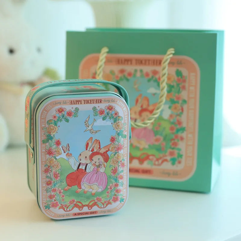 A decorative tinplate candy box with a yellow carrying loop is shown in front of a green gift box.