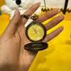 Eine Hand hält eine goldfarbene, vintage-Stil-Taschenuhr mit offenem Gehäuse und Kette vor gelbem Hintergrund