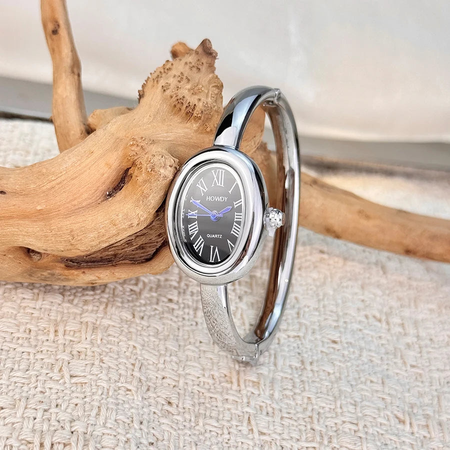 A silver quartz watch with a round face displaying Roman numerals and a metal band rests on a light fabric next to a piece of