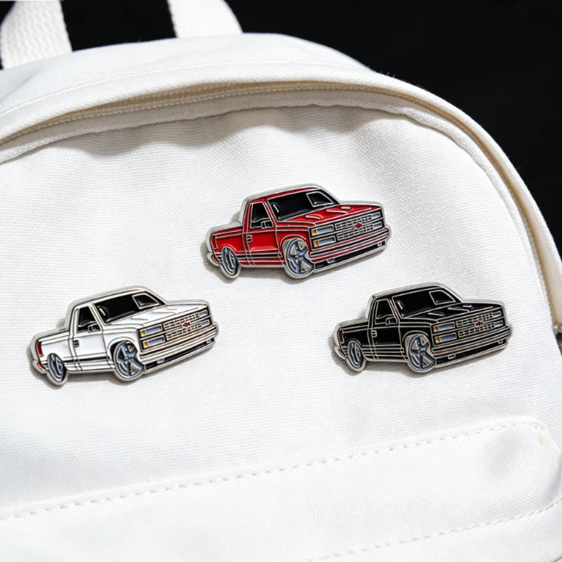 A white fabric pouch features three metal clip pins depicting classic American pickup trucks in red, silver, and black.