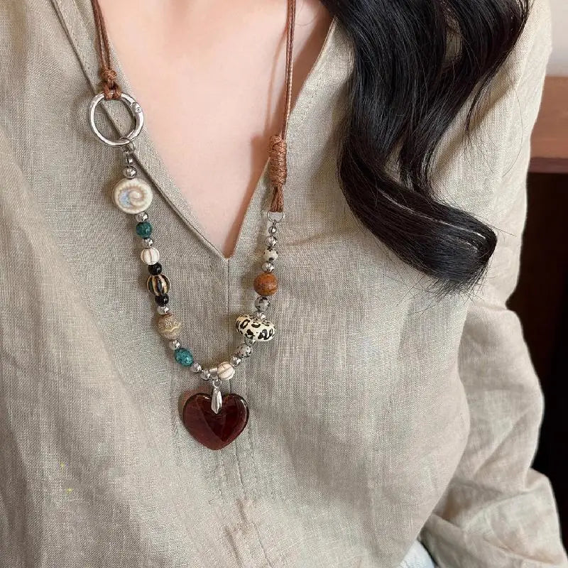 A close-up of a bohemian heart beaded pendant necklace worn against a light beige shirt.