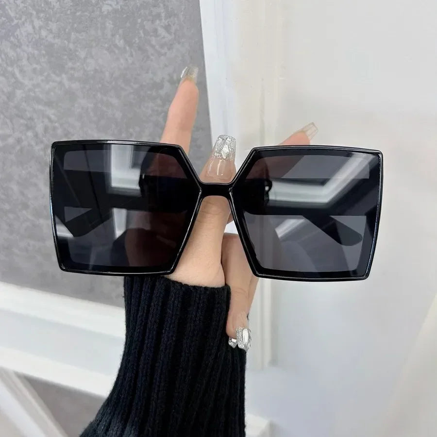 A hand wearing a black square mirror-finish sunglass frame is holding them up against a white background.