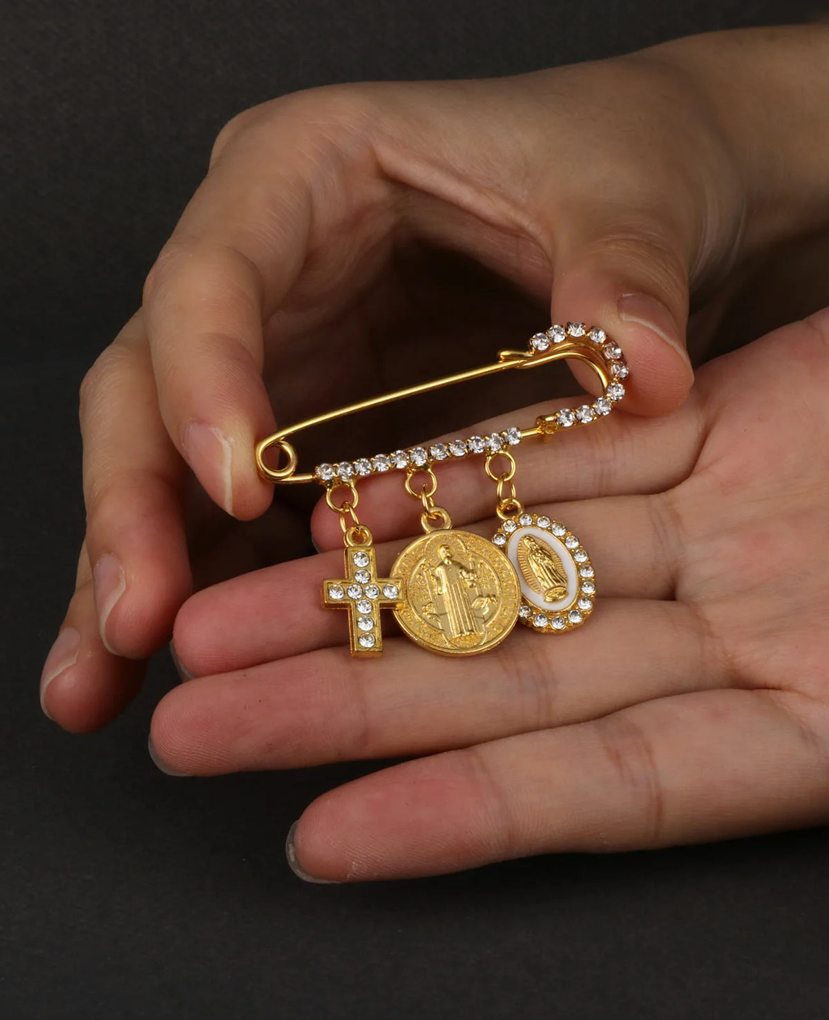 Eine Hand hält eine goldfarbene St. Benedikt-Medaille-Anstecknadel mit Kreuz und Figur auf einer Kette.
