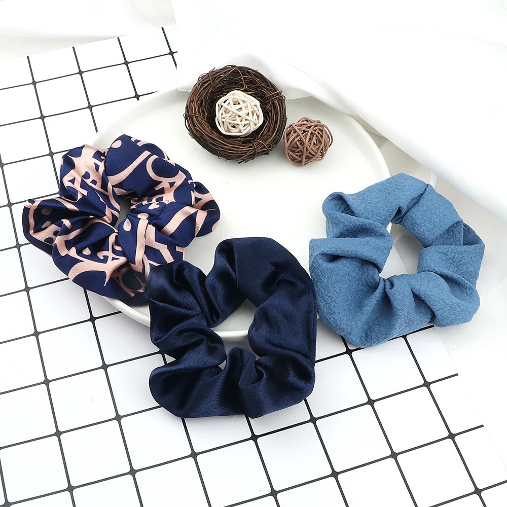 A set of three knitted hair scrunchie headbands in navy blue, dusty blue, and patterned fabric are displayed on a white surfa