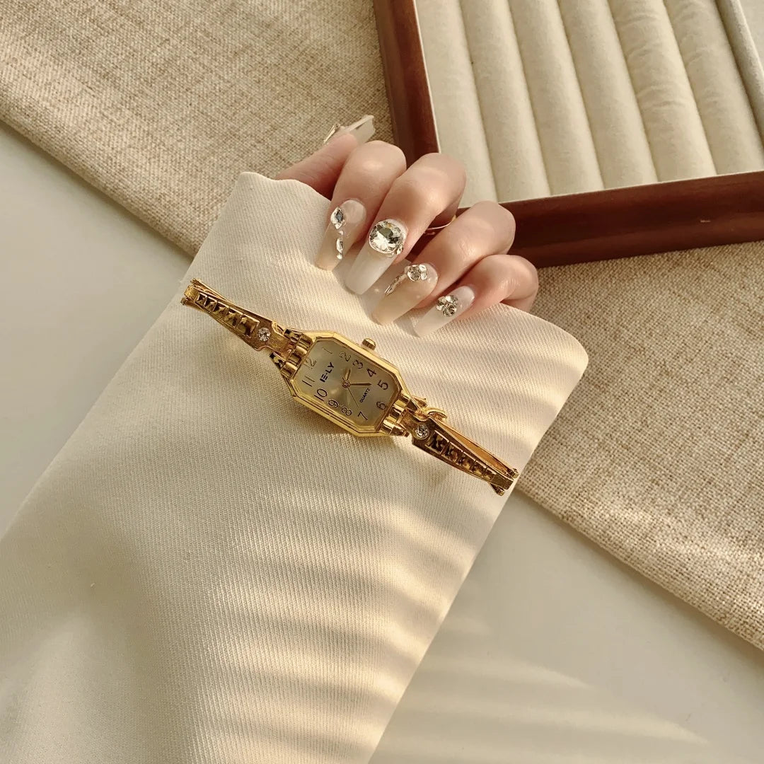 A close-up of a gold-toned quartz fashion wristwatch worn on a hand, resting on a beige fabric surface.