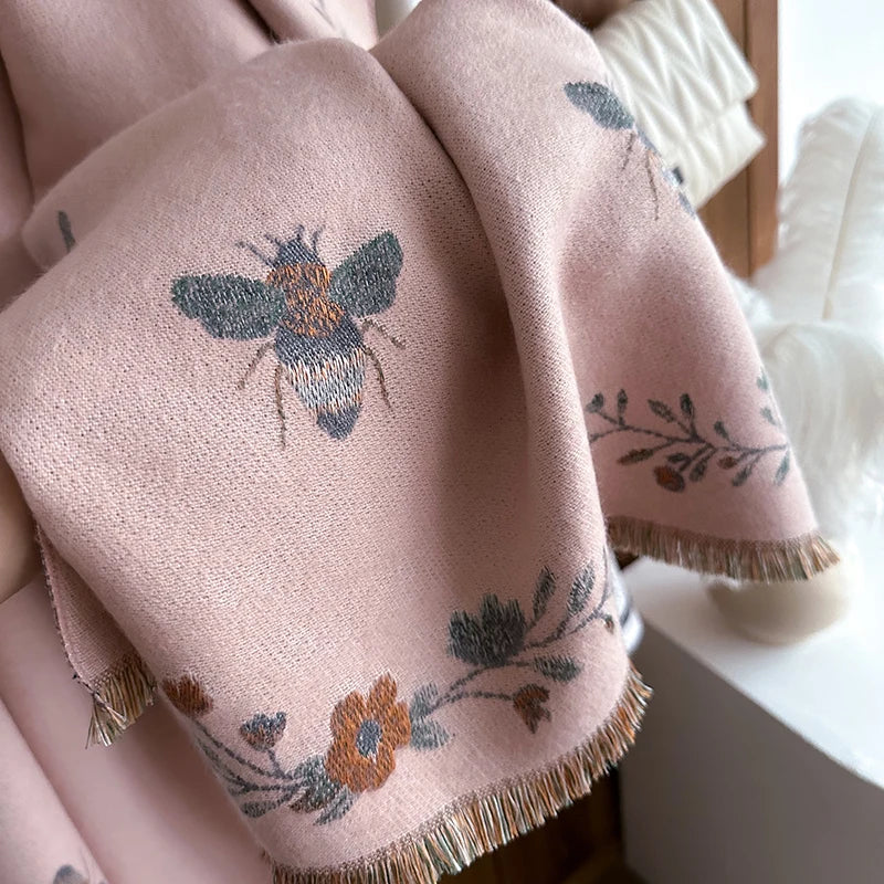 A soft pink pashmina shawl with a floral and bee print detail, featuring fringed edges.