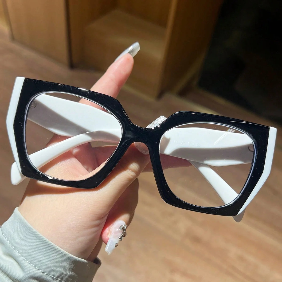 A hand holds a pair of black and white fashion glasses with large frames against a blurred background.