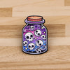 A dark purple enamel pin featuring multiple white skulls and swirling patterns, set in a glass-like container.