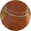 Layered gold-plated twisted ankle bracelets displayed on a brown skin tone.