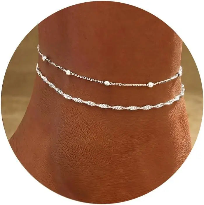 Layered gold-plated twisted ankle bracelets displayed on a brown skin tone.