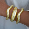 A set of three gold geometric wave bangles worn on a wrist.