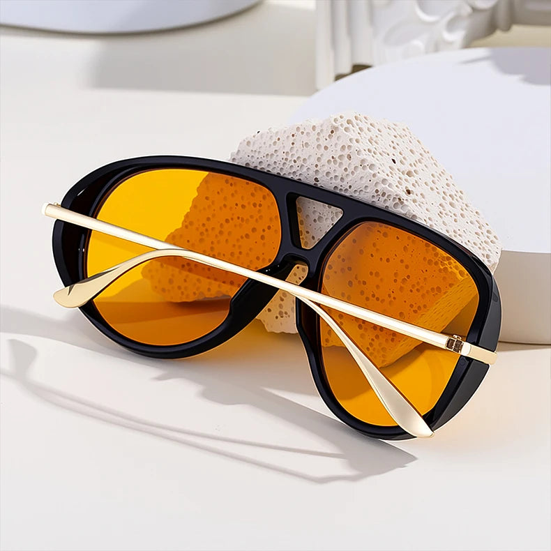 Un par de gafas de piloto vintage grandes con montura negra y lentes ámbar sobre una superficie blanca junto a una texturizad
