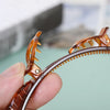 A hand is holding a multi-layer braided hair headband clip, showing the detailed texture and orange-brown color.