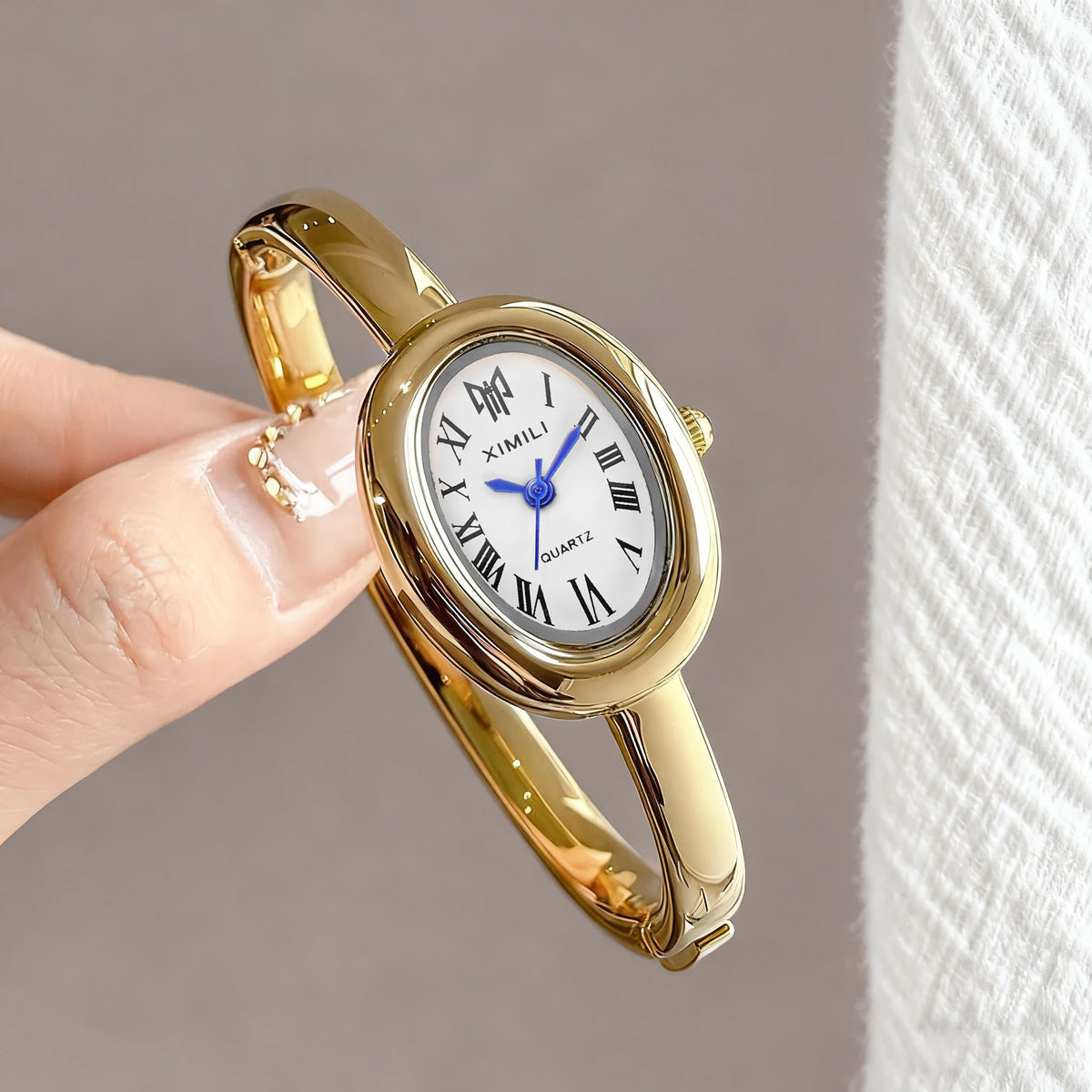 A close-up shot of a gold-toned quartz watch bracelet with a white face and blue hands being held by a person's fingers.