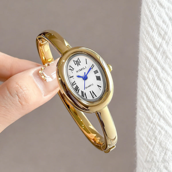 A close-up shot of a gold-toned quartz watch bracelet with a white face and blue hands being held by a person's fingers.