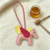 A pink horse charm with yellow accents hangs from a pink cord next to a small glass jar of honey on a cream fabric background