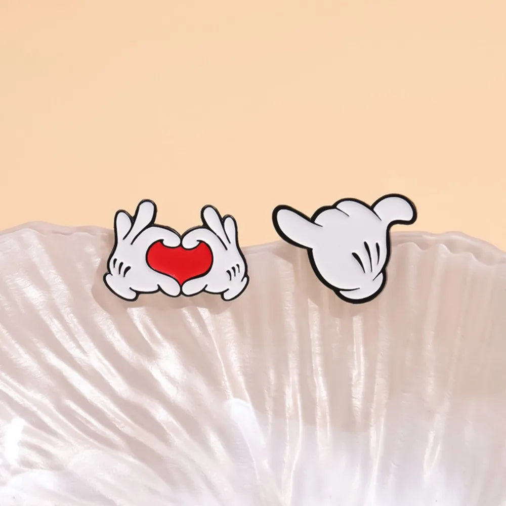 A pair of enamel pins depicting cartoon hands forming a heart shape and a thumbs-up gesture on a white background.