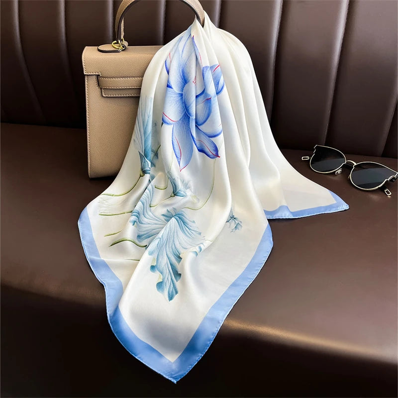 A white silk scarf with blue floral and leaf patterns is draped on a brown leather sofa next to a beige clutch and sunglasses