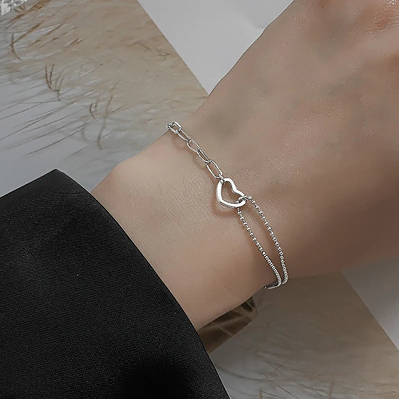 A close-up of a sterling silver double heart charm bracelet worn on a wrist against a dark sleeve.