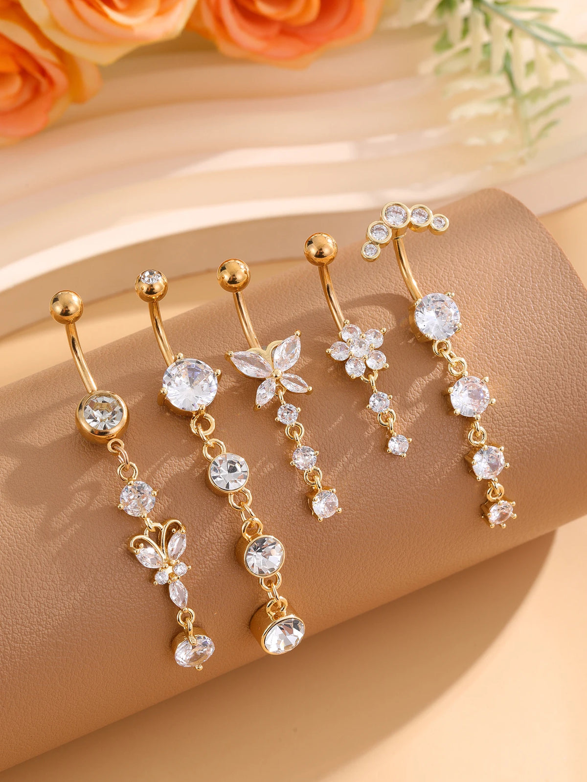 A set of five gold stainless steel dangled belly button rings with clear and white stones displayed on a beige surface.