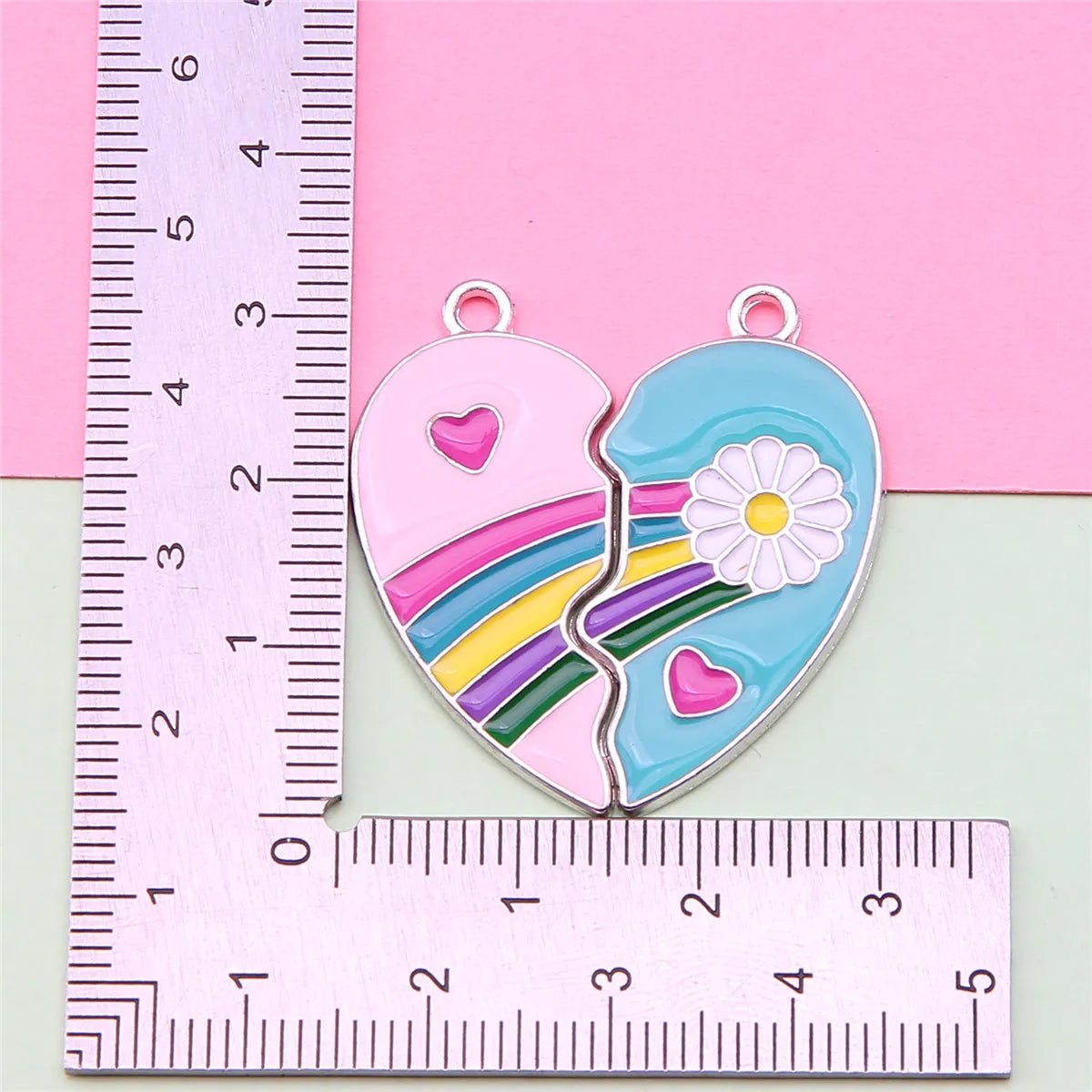 Dos colgantes de collares de amistad en forma de corazón, uno rosa y uno azul, mostrados junto a una regla.