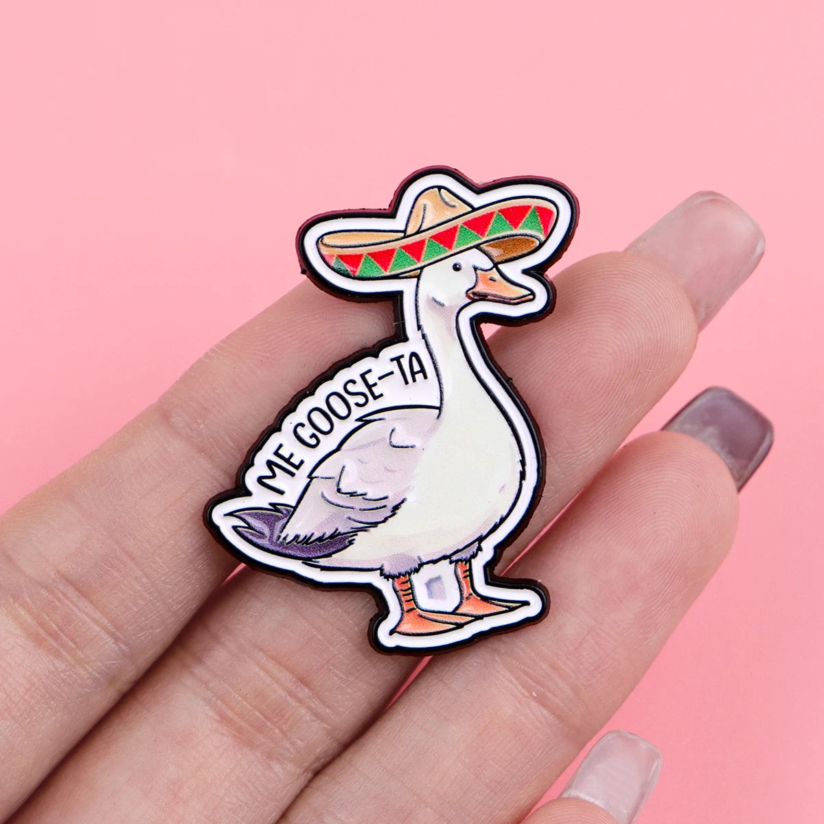 A hand holds a enamel pin featuring a goose wearing a sombrero and the text ME GOOSE-TA