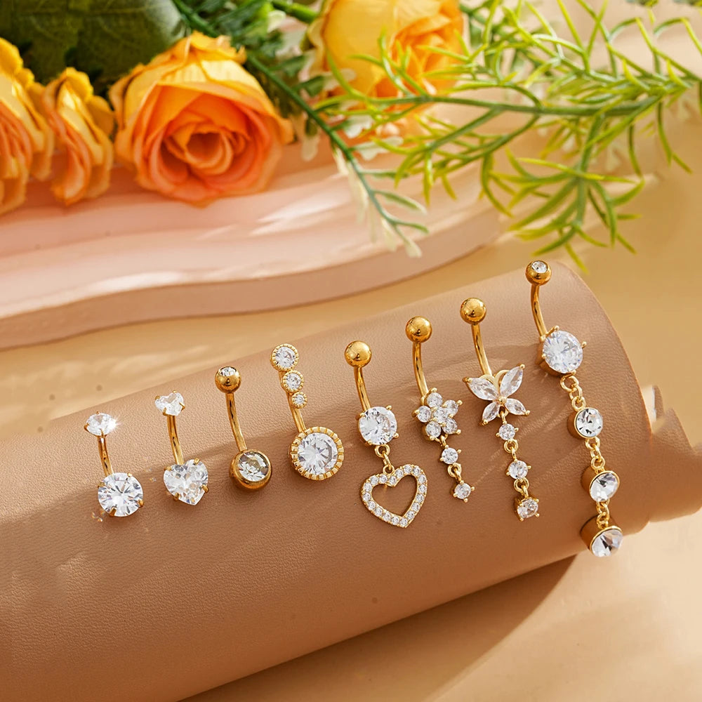 A set of gold dangle curved belly piercing jewelry featuring heart and flower designs displayed on a beige surface.